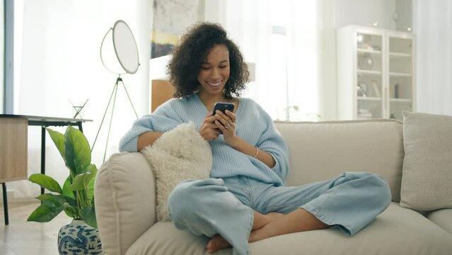 Bi racial girl uses mobile phone watching social media sitting at home in sunlight. Real people lifestyle smartphone entertainment 4K. Smiling afro american enthicity woman holding in hands smartphone