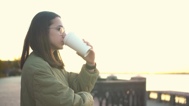 Young Woman With A Glass Of Coffee In Her Hand. A Girl In Glasses Near The Fence On The Embankment With A Hot Drink In A Glass.