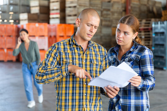 Man and young woman talking about documentation while standing in warehouse. Another woman talkin on phone in background.