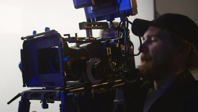 Close-up shot of Caucasian male videographer holding video camera or camcorder on shoulder mount, giving instructions to assistant, who is adjusting settings, while getting ready for filming in studio
