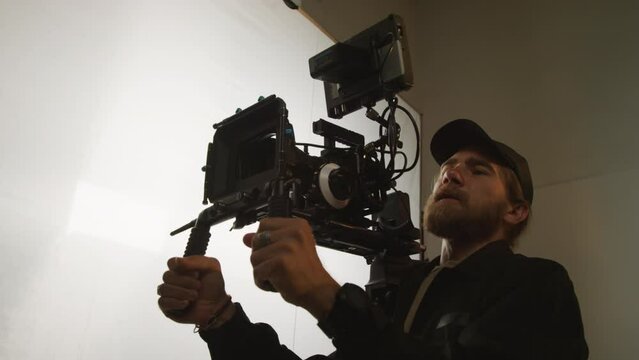 Medium close-up shot of Caucasian cameraman holding professional video camera on shoulder mount with stabilizer, filming fashion commercial and giving directions to models in frame
