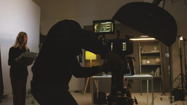 Medium shot of production crew preparing for commercial shoot in studio - videographer installing camera on tripod, female producer and assistant discussing storyboard, presenter looking through text