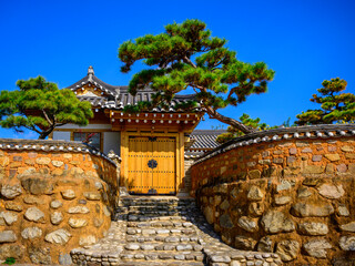 Korean traditional house, stone and mud wall, and green curved pine trees against the blue sky...