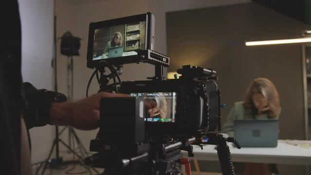 Close-up shot of hands of cameraman preparing equipment for shooting online show and setting up frame on camera touchscreen, while female presenter with laptop is preparing material