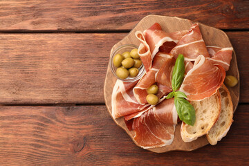 Slices of tasty cured ham, olives, bread and basil on wooden table, top view. Space for text