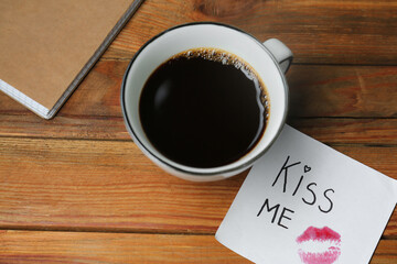 Sticky note with phrase Kiss Me, lipstick mark and cup of coffee on wooden table, above view