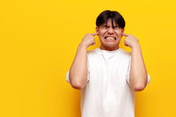 young asian guy in white t-shirt covers ears with hands and ignores loud sound on yellow isolated background - obrazy, fototapety, plakaty