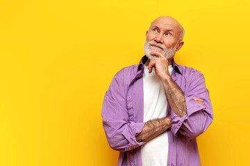 pensive old bald grandfather with gray beard in purple shirt stands with his arms crossed on yellow...