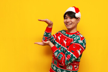 young asian man in christmas sweater and santa hat holding empty hands on yellow isolated background