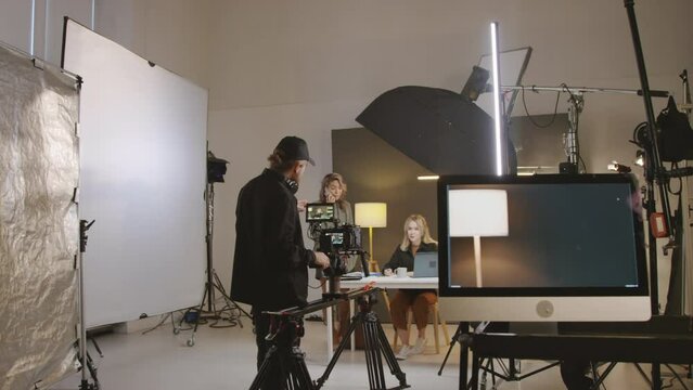 Full shot of production crew preparing for video shoot in studio - operator standing next to video camera on tripod, female producer talking on mobile, presenter sitting at desk, discussing script