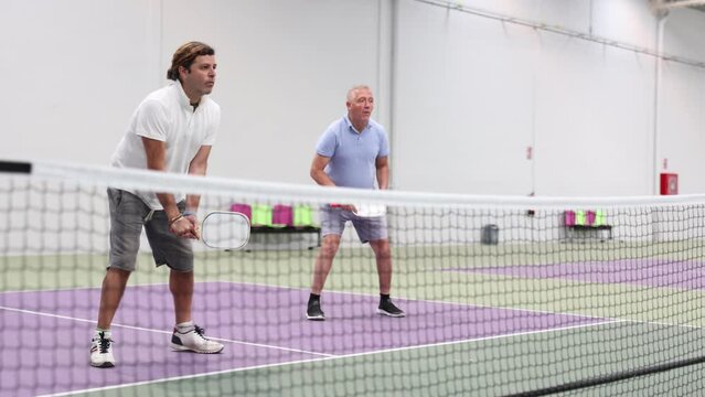 Portrait of concentrated adult pickleball player preparing to strike and return ball to opponents field during doubles match in team with older partner 
