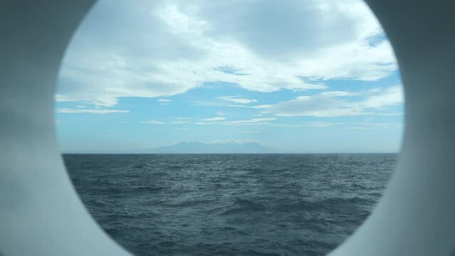 View from round deck window to sea. Clip. Beautiful seascape with mountain coast on horizon from round window. Sea voyage and view from round window