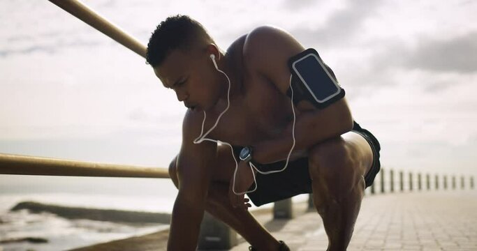 Runner man, tired and earphones by ocean promenade fence for breathing, fitness or listening to music. African person, training and exercise with fatigue, thinking or phone for streaming subscription