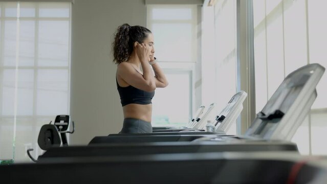 Side view of beautiful muscular woman looking out of window and speaking with a friend using wireless earphones during conversation, doing exercises in the morning at a gym.