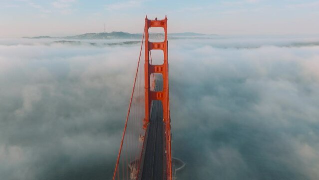 Karl the Fog drifting below red bridge main landmark of San Francisco slow motion California USA. Aerial video cinematic Golden Gate Bridge. Cars driving by red bridge covered in scenic white clouds