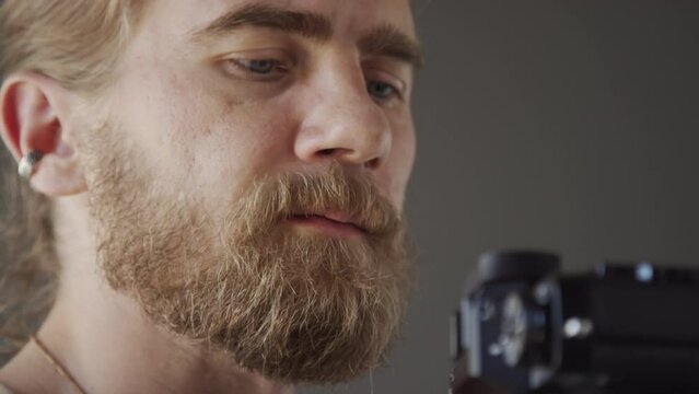 Close-up shot of hands and face of busy Caucasian male photographer with red hair and beard taking flatlay photos with camera on tripod, pressing button the adjusting focus