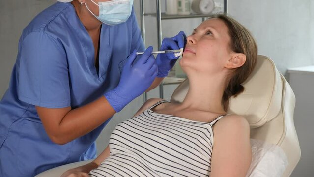 Unrecognizable circumcised female cropped doctor in disposable medical mask manipulates introduction of filler and correction of shape and size of lips to girl patient. Close-up