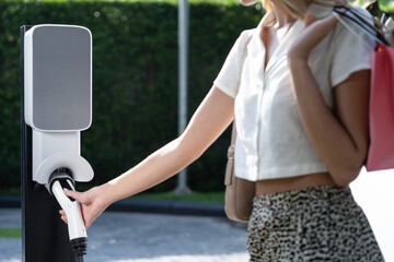Young woman travel with EV electric car charging in green sustainable city outdoor garden in...