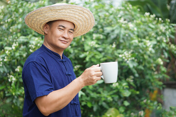 Asian man farmer wears hat, blue shirt, holds cup of coffee in garden. Concept, relax time with...