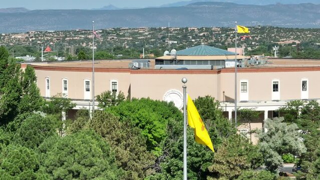 New Mexico state flag waving in front of capitol building in Santa Fe, NM. Aerial establishing shot on bright day.