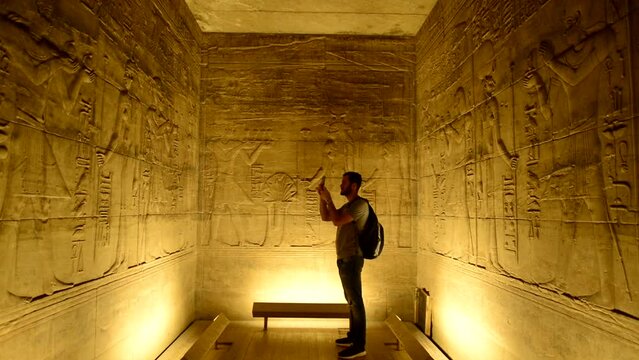 Tourist man photographing a room of egyptian temple of Horus in Edfu. Traveler with a bag in egypt, visiting Horus Temple in Edfu, Egypt. Explorer, tourist takes photos of hieroglyphs on Egypt walls
