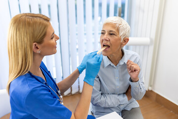 Senior patient opening her mouth for the doctor to look in her throat. Female doctor examining sore throat of patient in clinic. Otolaryngologist examines sore throat of patient.