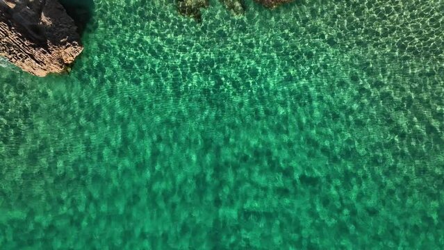 Top down drone shot of shallow crystal clear water towards a rocky coastline