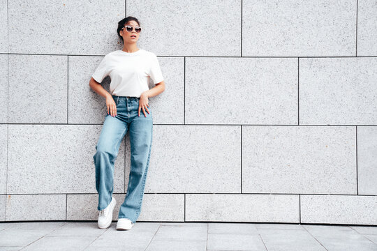 Young beautiful smiling hipster woman in trendy summer white t-shirt and jeans clothes. Carefree woman, posing in the street at sunny day. Positive model outdoors near wall. In sunglasses