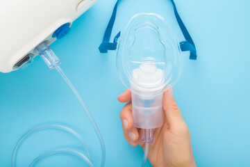 Fototapeta premium Young adult woman hand holding mask with container for drug breathing. Compressor nebulizer on light blue table background. Point of view shot. Closeup. Medical device for inhalation. Top down view.