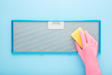 Woman hand in pink rubber protective glove cleaning kitchen cooker hood filter with yellow sponge on light blue table background. Pastel color. Closeup. Top down view. - obrazy, fototapety, plakaty