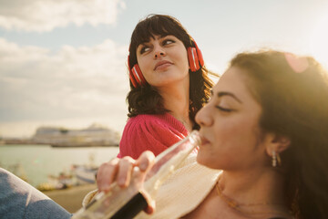 Two women savor a tranquil moment by the sea. One with headphones gazes into the distance, while...