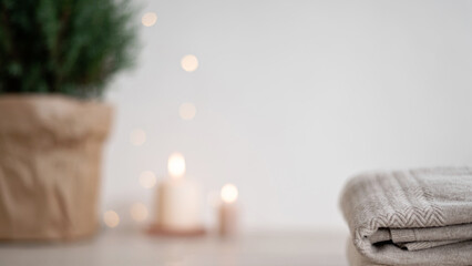 Blurred defocused background with candle lights, pine in pot and empty white wall. Wool winter sweater on table on foreground. Cozy white Christmas home interior backdrop, banner