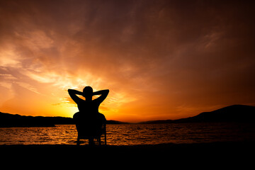 sylwetka osoby o zachodzie słońca siedząca i podziwiająca zachód słońca, relaks nad morzem - obrazy, fototapety, plakaty