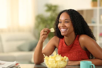 Gordijnen Happy black woman eating potato chips at home © Antonioguillem