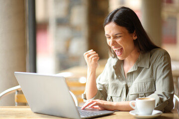 Excited woman celebrating in a restaurant checking laptop - obrazy, fototapety, plakaty
