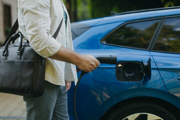 Close up of businessman charging his electric car in front of house. Pluging charger in charging port.