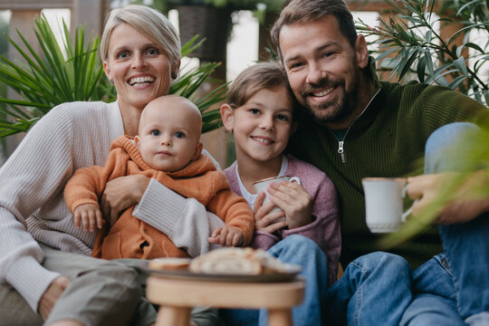 Portrait of family sitting outdoors in the garden patio. Drinking warm tea, coffee, and eating sweet roll bread. Hygge family autumn time. - Powered by Adobe