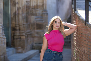 Young woman, blonde, green eyes, wearing pink t-shirt and denim skirt, looking at camera, pensive and calm, leaning against a brick wall. Concept beauty, thoughts, dreams, tranquility.