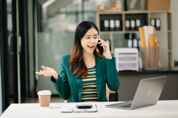 Asian Business woman Talking on the phone and using a laptop with a smile while sitting at office