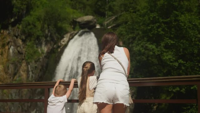Little children spellbound by waterfall and mother turns back on park bridge. Woman with cute kids travel to look at river cascade at wilderness resort