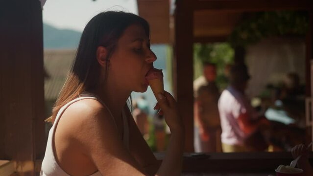Languid woman bites tasty ice cream in wafer cone sitting at table in cafe slow motion. Lady enjoys frozen dessert resting on restaurant terrace