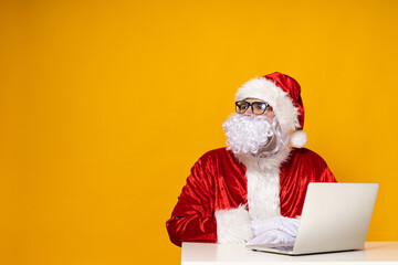 Santa Claus, at the table with a laptop, on a yellow background.