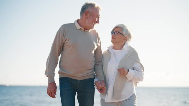 Relaxed sensual senior couple wife husband walking on sea beach enjoying time talking holding hands in sunny evening. Loving family man woman talking. Lifelong relationships, happy marriage concept.