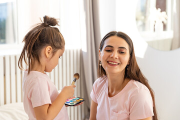 Smiling mother and preschool daughter doing makeup together, excited little girl, holding makeup brush, applies powder to mother's face, happy child applying blush, having fun playing with mom at home
