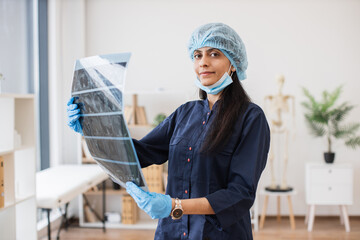 Portrait of hindu medical worker dressed in dark blue uniform and disposable cap holding result of x-ray scan of sick patient in cabinet. Competent doctor investigating diseases before surgery.