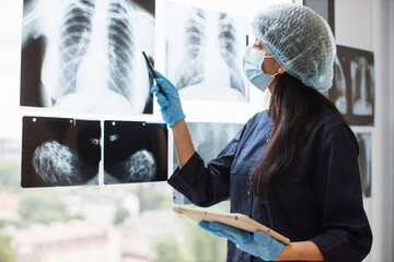 Focused female medical worker in face mask and scrub top posing with digital tablet near x-ray scans fixed on window glass. Qualified hindu surgeon in medical gloves studying injuries.