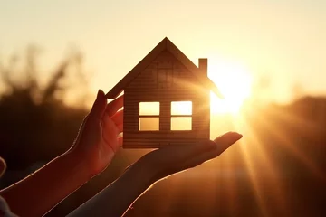 Foto op Canvas A symbol of your cozy home against the backdrop of the setting sun. Little wooden house in female hand delicately conveying dreams and hopes for secure. © Stavros