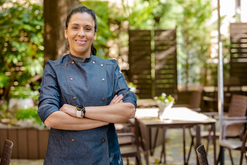 Female chef wears blue coat, happy smiling poses with arms crossed outdoors on the terrace of a restaurant. Restaurant worker, culinary gourmet, pastry chef. - obrazy, fototapety, plakaty