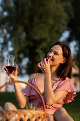 Happy young woman sitting on blanket eating grape and drinks wine outdoors. Beautiful girlfriend enjoying picnic lunch in park. Caucasian lady drinking alcohol at the garden