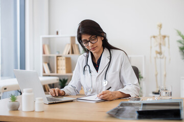 Concentrated female doctor with stethoscope sitting at table and writing prescription while using modern laptop. Indian nurse looking at information about medicine and taking notes in medical office.